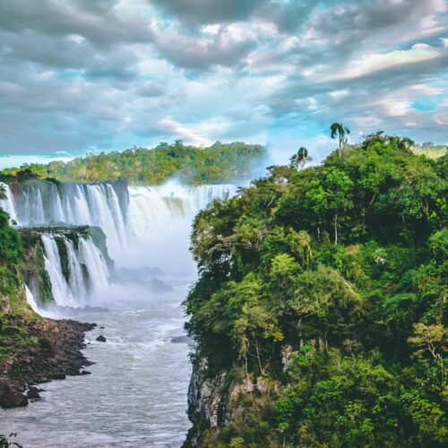 Breathtaking view of towering waterfalls surrounded by vibrant green jungle and dramatic clouds.